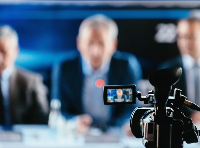 camcorder in focus aimed at three men, out of focus, dressed in suits and sitting at desk with papers and water bottles