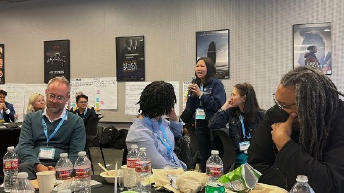 Emily Eng standing up, holding microphone and speaking to an audience seated at tables.