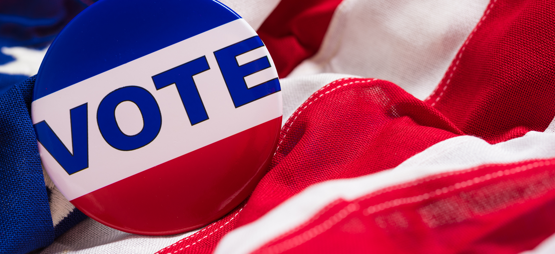 Button pin with word "VOTE" resting on a rumpled American flag.