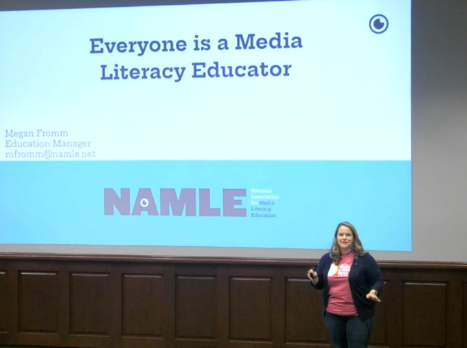 Speaker from the organization NAMLE faces audience of educators, standing in front of a screen with a slide that reads "Everyone is a Media Literacy Educator, NAMLE."