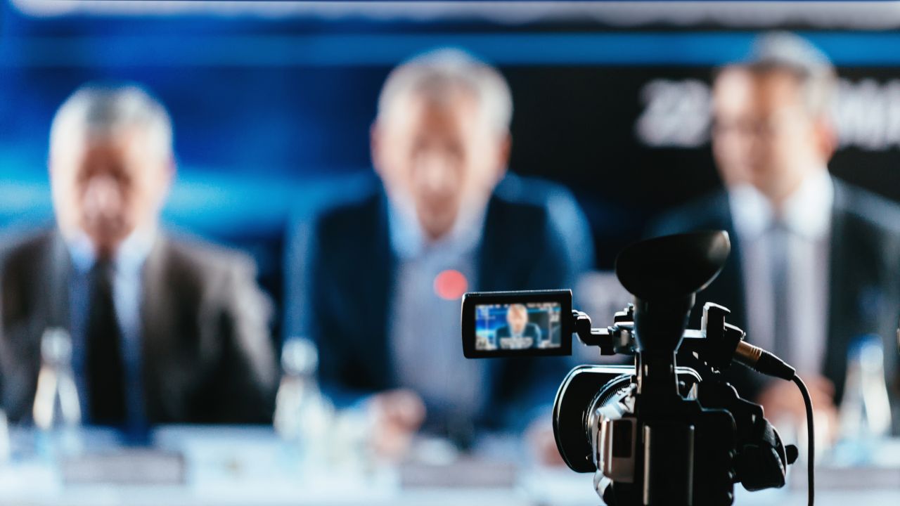 camcorder in focus aimed at three men, out of focus, dressed in suits and sitting at desk with papers and water bottles