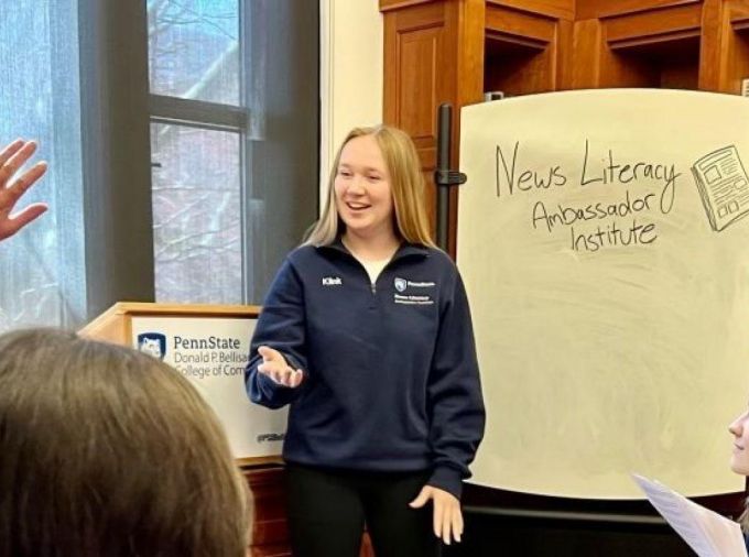 person standing and facing three other people, in front of a easel with writing that says "news Literacy Ambassador Institute" and bookshelves. One person is raising their hand.