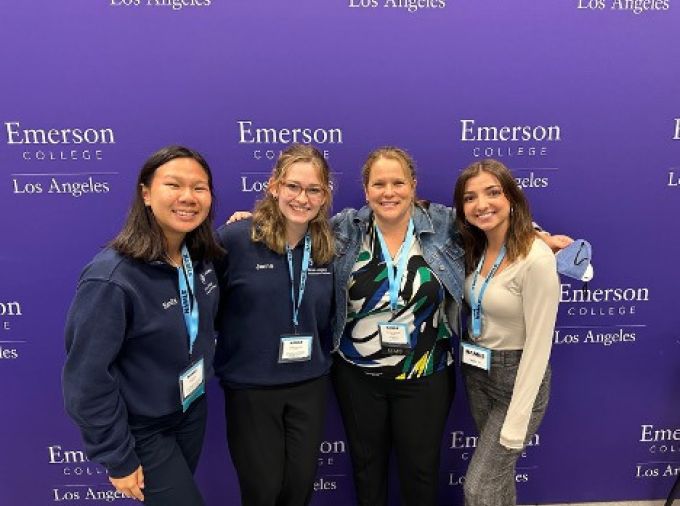 four women with name tags on lanyards, pose with arms around each other in front of a purple backdrop that reads "Emerson College Los Angeles"