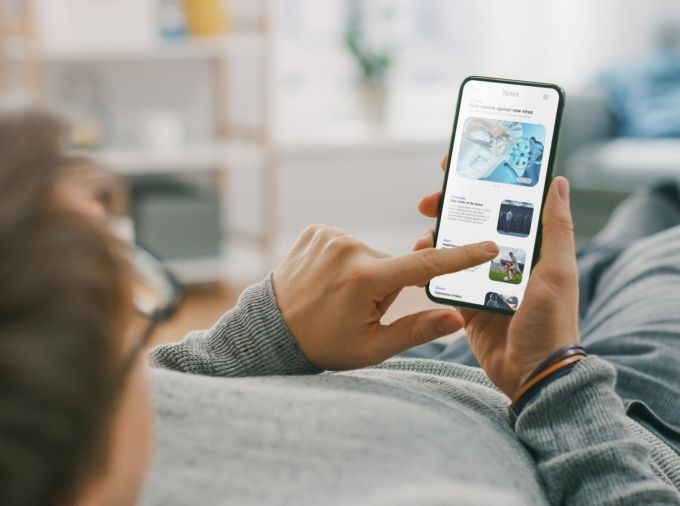 A man on a couch scrolling through news articles on a smartphone